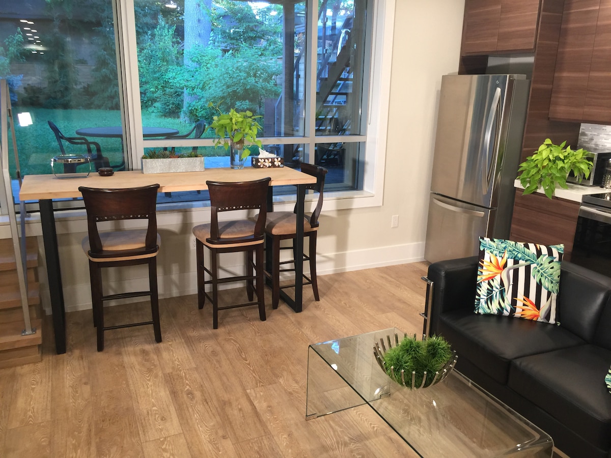 A modern kitchen area features a high wooden table with three bar stools, positioned beside a large window that offers views of the backyard greenery. A sleek silver refrigerator stands nearby, and a black leather couch is visible in the living space, complemented by a decorative plant arrangement on a glass table.