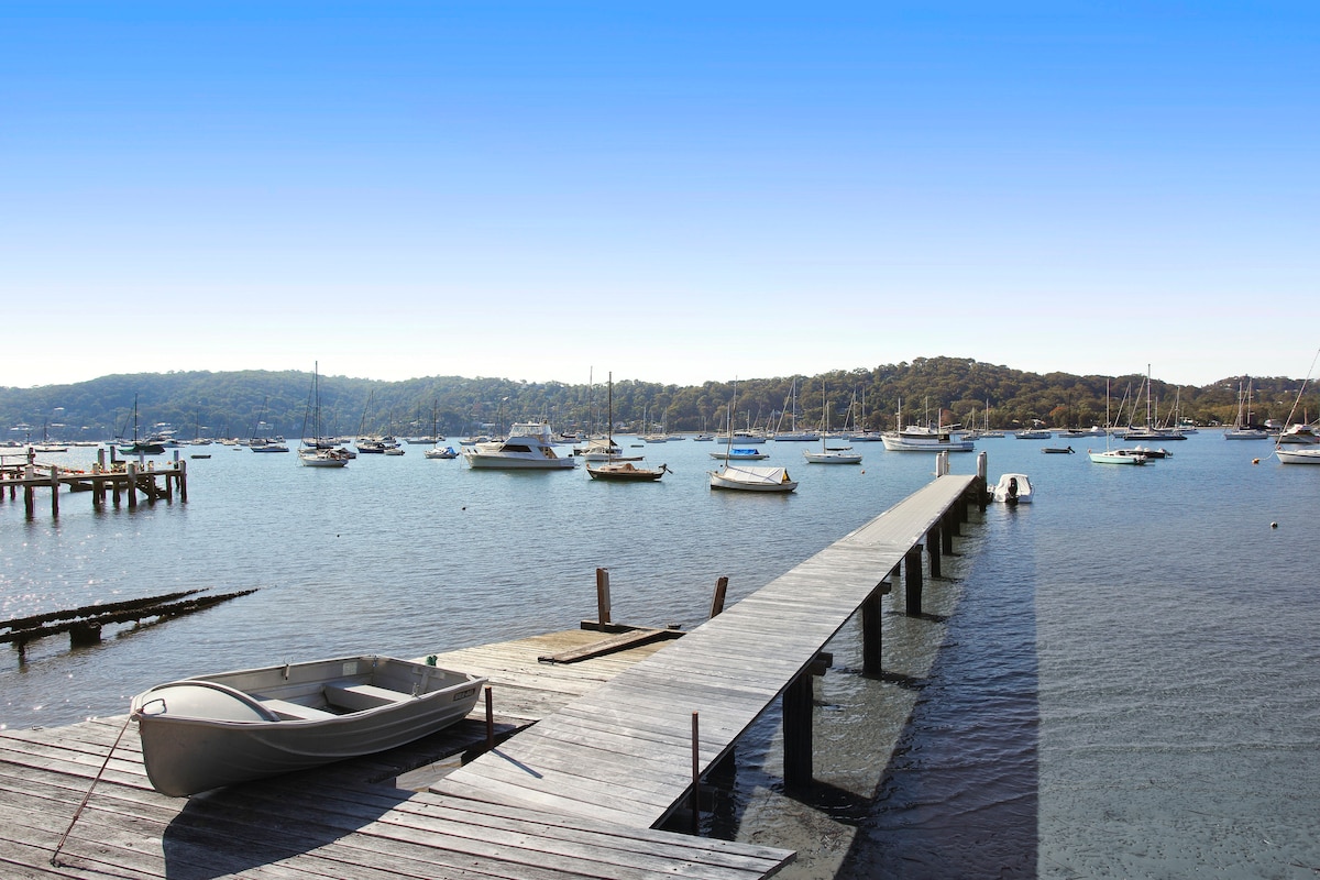 A wooden jetty extends into Careel Bay, lined with boats resting on the calm waters. The tranquil landscape features rolling hills in the background, creating a serene atmosphere. Clear blue skies reflect on the water's surface, enhancing the peaceful waterfront setting.