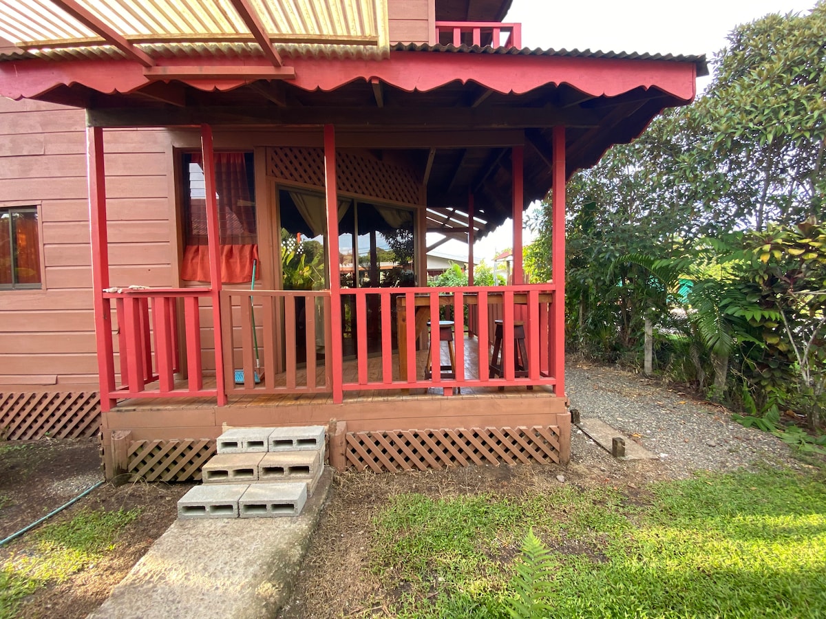 The exterior of the cabin features a covered porch with red railings and a wooden deck. Steps lead up to the entrance, and the area is surrounded by green foliage, creating a natural, inviting space for relaxation.