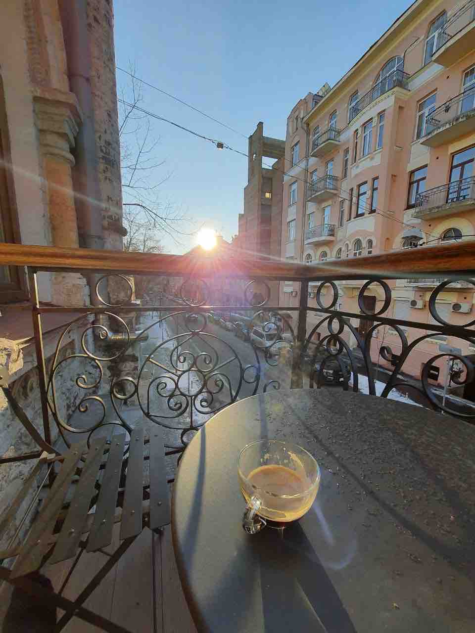 A wrought-iron balcony rail frames a small table adorned with a glass of coffee. The sun rises in the background, casting a warm light over the street below, accentuating the architectural details of the surrounding buildings.
