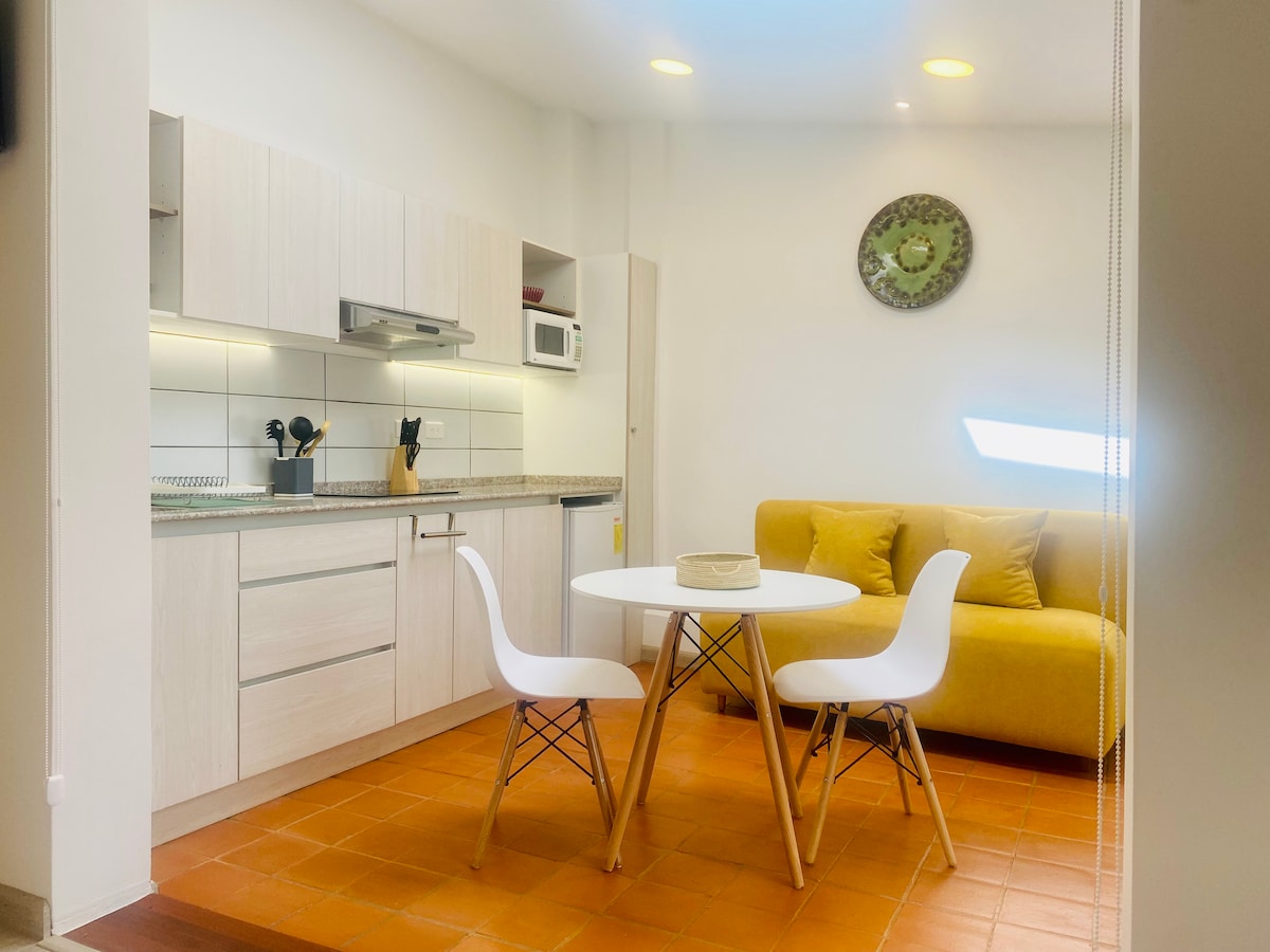 A bright kitchen area is presented with modern cabinetry and a countertop. A round dining table for two is surrounded by white chairs. A yellow sofa adds a pop of color, while natural light from a nearby window enhances the space.