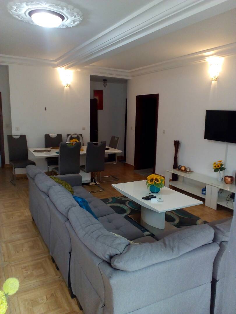 A modern living space is highlighted by a light gray sectional sofa facing a white coffee table. A vibrant area rug adds color beneath the furniture. The dining area, visible in the background, features a table set for six, complemented by stylish gray chairs.
