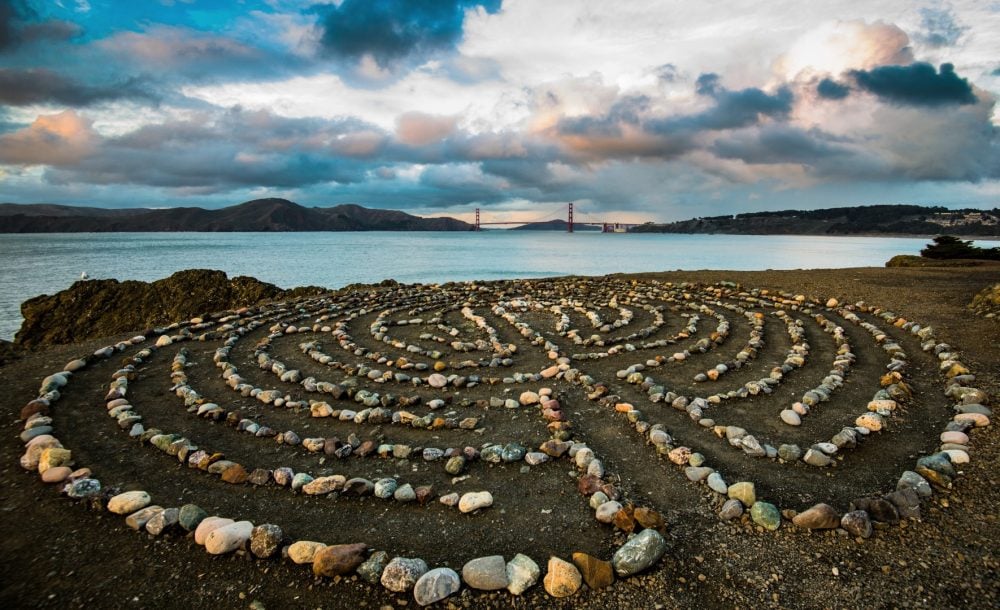A spiral labyrinth made of smooth stones is situated on a sandy shore. The serene waters of the bay extend in the background, with the Golden Gate Bridge visible across the horizon under a cloudy sky. Natural textures and earthy tones dominate the scene.