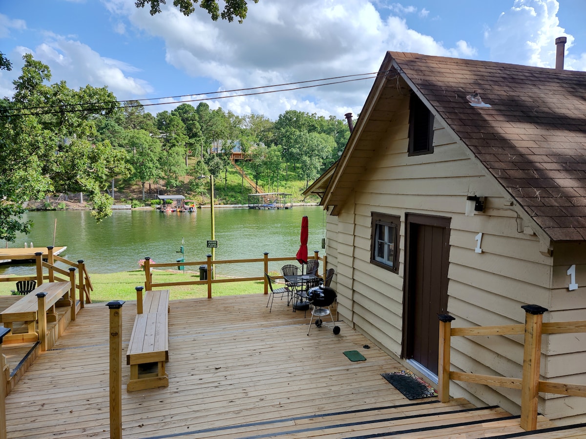 The outdoor deck features wooden seating and a grill, providing a space for relaxation. A view of the water and tree line enhances the setting. The cottage is situated nearby, with a door and window visible, framed by sunny skies and scattered clouds.