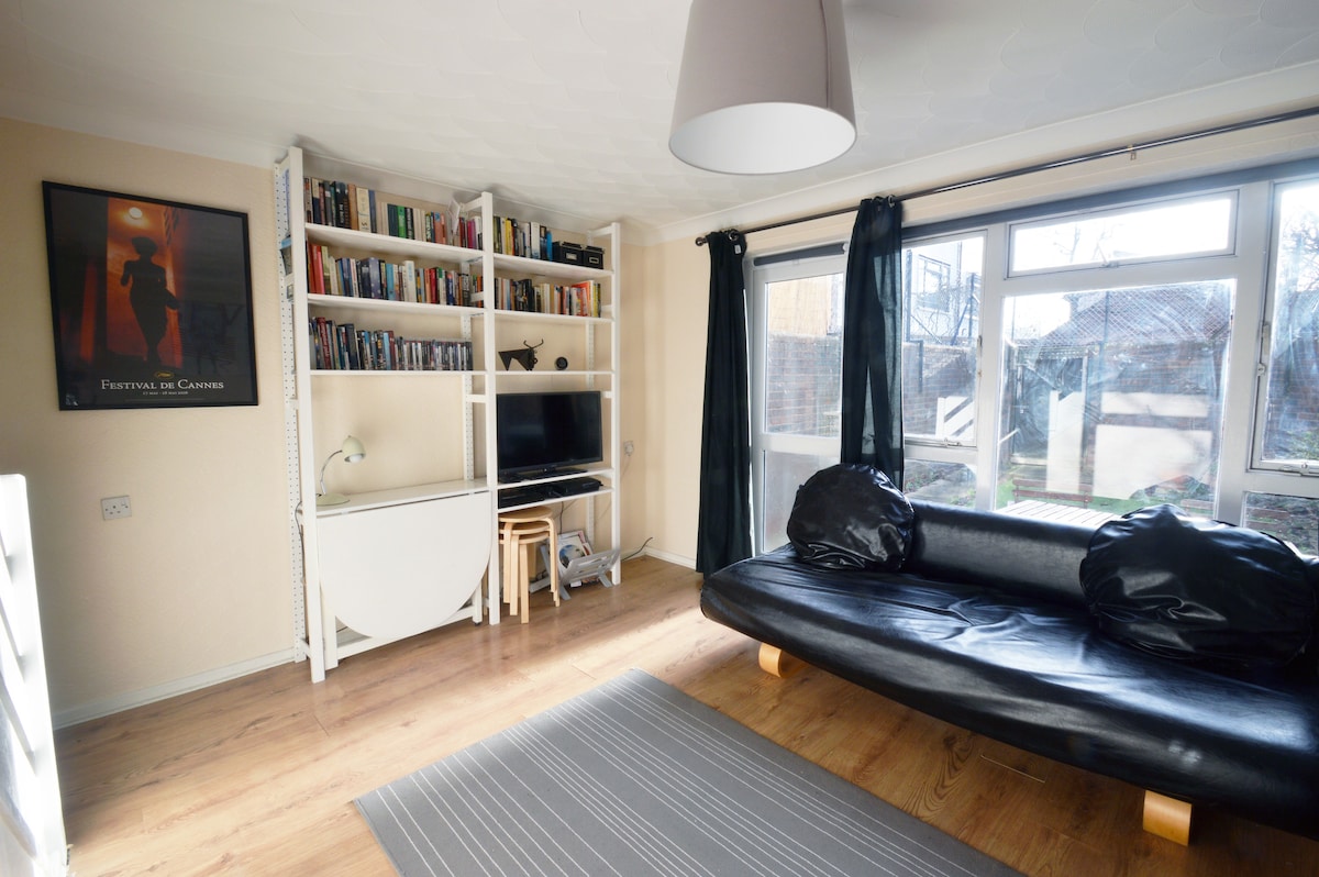 The lounge area features a black leather sofa, a small dining table with chairs, and a TV on a modern stand. A large window allows natural light to illuminate the space, which includes built-in bookshelves filled with books. The floor is covered with a striped rug.