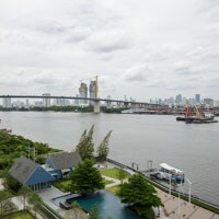 A serene view of the Chao Phraya River is captured, showcasing green spaces alongside the water. The modern skyline is visible in the background, featuring notable structures and a bridge that spans across the river. Calm waters reflect the surrounding landscape.
