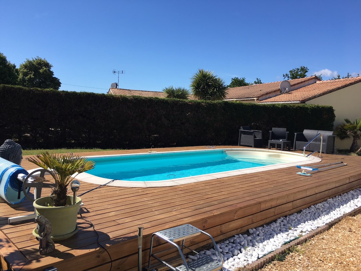 An outdoor swimming pool is situated on a wooden deck surrounded by neatly trimmed hedges. Comfortable seating arrangements are placed nearby, complemented by potted plants that add greenery. A bright blue sky enhances the serene atmosphere of the outdoor area.