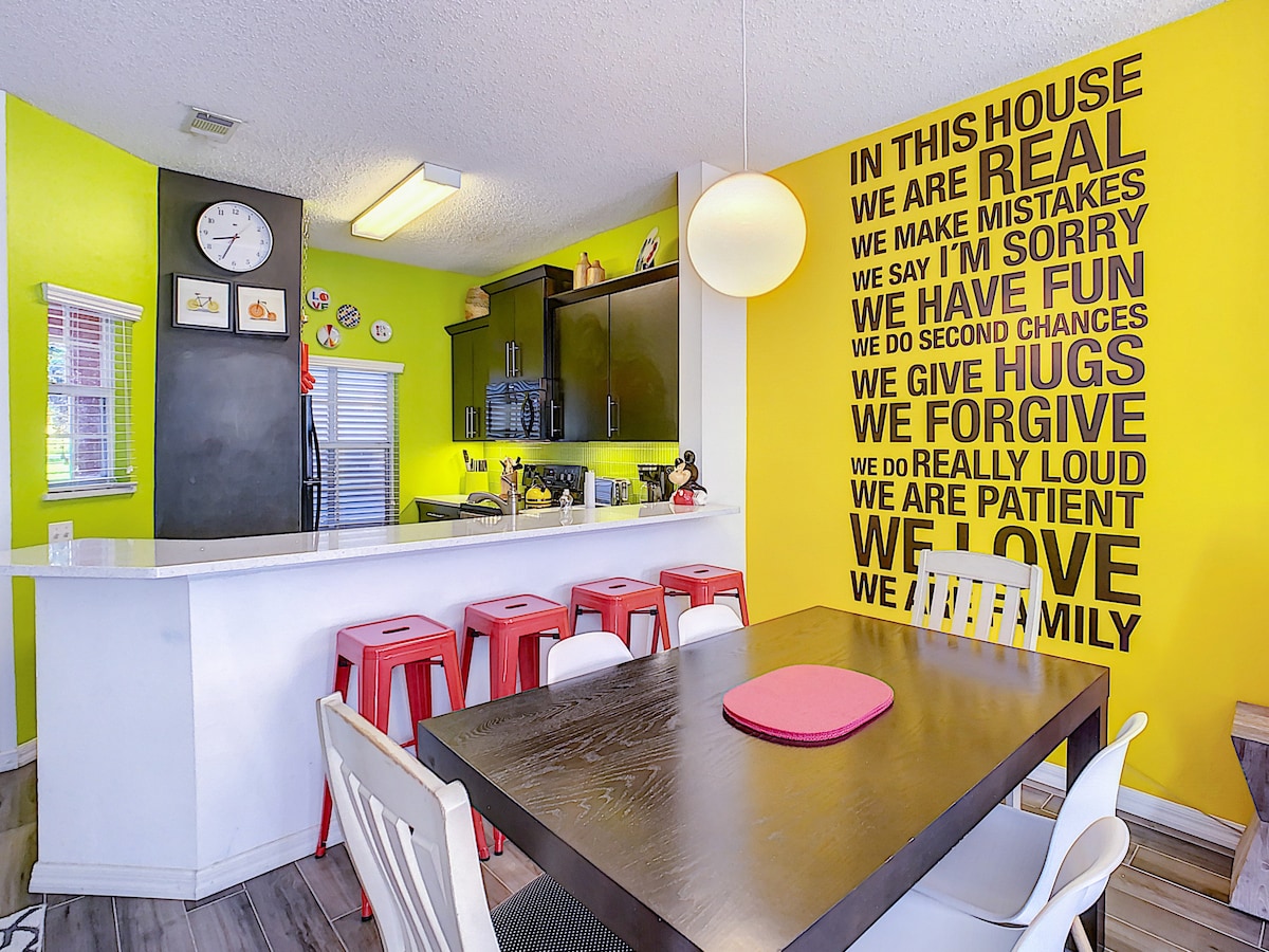 A vibrant kitchen space features a large dining table surrounded by white and red chairs. A bright yellow wall displays inspirational text. The kitchen island displays modern bar stools, while black cabinets and counter spaces complement the lively atmosphere.