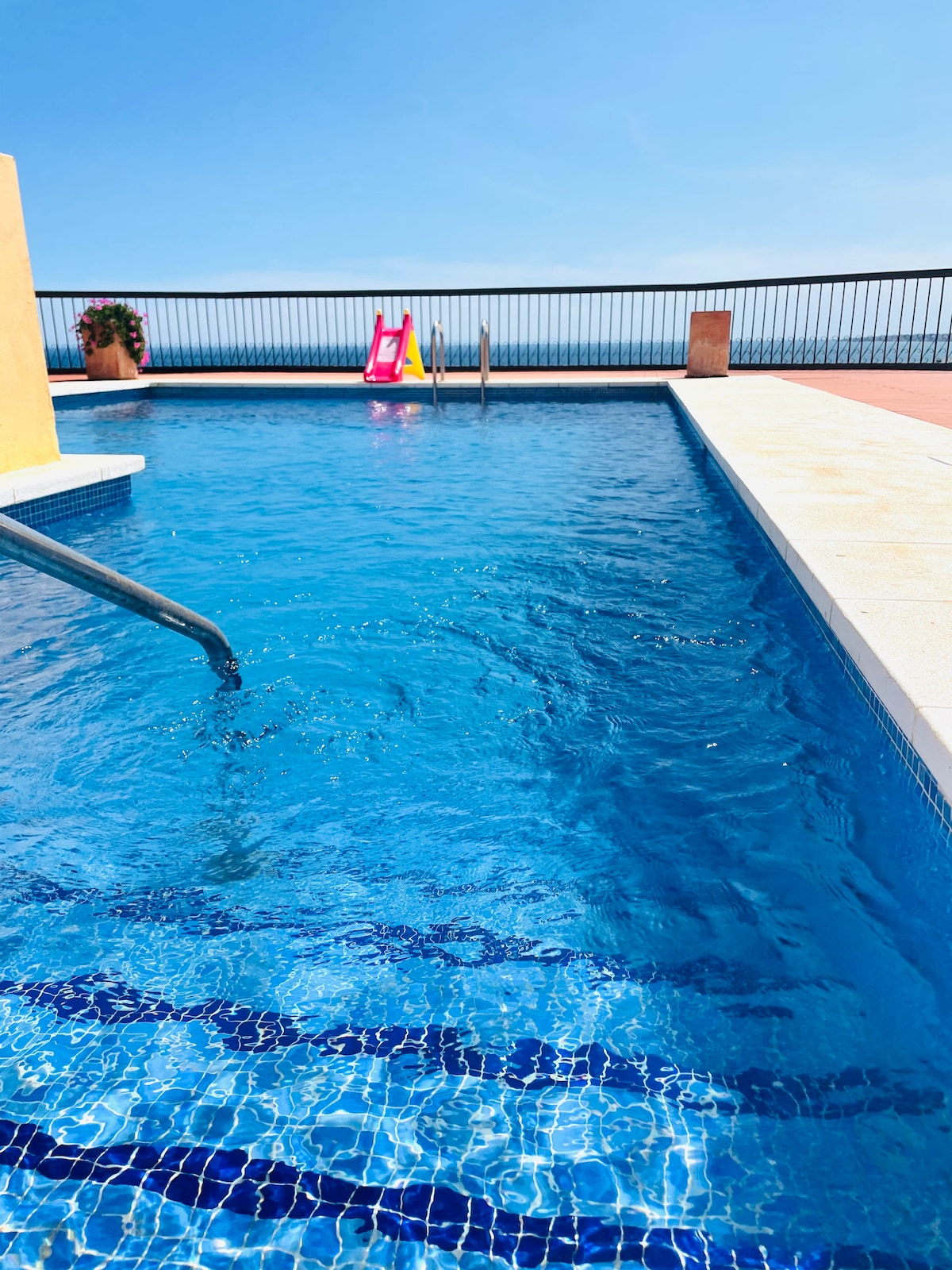 A sparkling pool is featured, with clear blue water reflecting the sky. Steps lead into the pool, and a colorful slide can be seen alongside the edge. The area is enclosed by a metal railing, offering views of the ocean in the background.