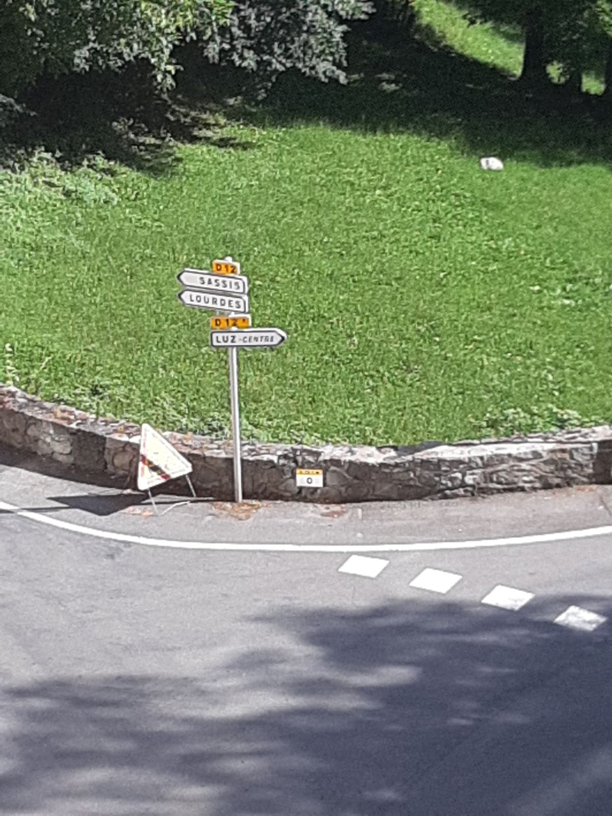 A street sign is positioned at an intersection, prominently displaying directions to nearby locations including Luz-Center and Lourdes. A grassy area with trees surrounds the sign, while a caution sign rests on the ground nearby, emphasizing the roadside setting.