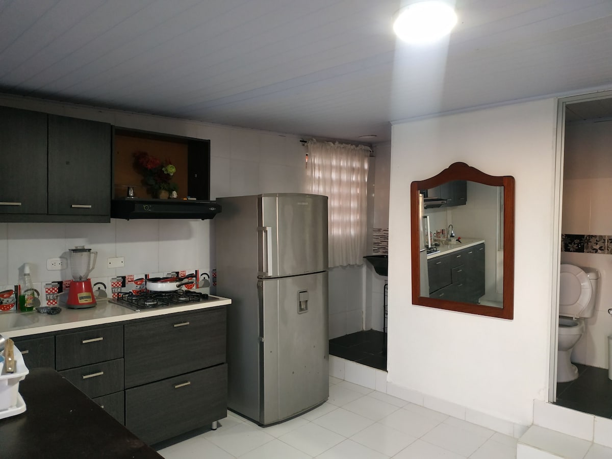 The kitchen area features modern cabinetry in dark wood, complemented by a stainless steel refrigerator and a gas stove. A window allows natural light, enhancing the space. A mirror above a dark wood cabinet reflects the room, while basic kitchen amenities are visible on the countertop.