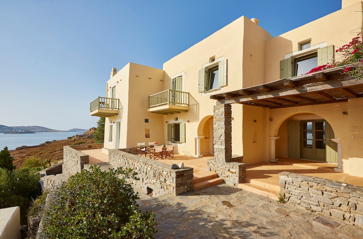 A spacious villa exterior is showcased, featuring two balconies with green shutters. The stone structure is complemented by a terrace area, where wooden chairs and a table are arranged. Surrounding greenery and a view of the ocean enhance the serene atmosphere.