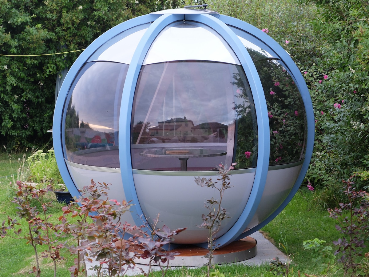 A modern spherical structure sits in a grassy area, surrounded by greenery. Large glass panels form the walls, allowing views of the landscape. A table is visible inside, providing a cozy spot for relaxation or dining. The design adds a unique touch to the outdoor space.