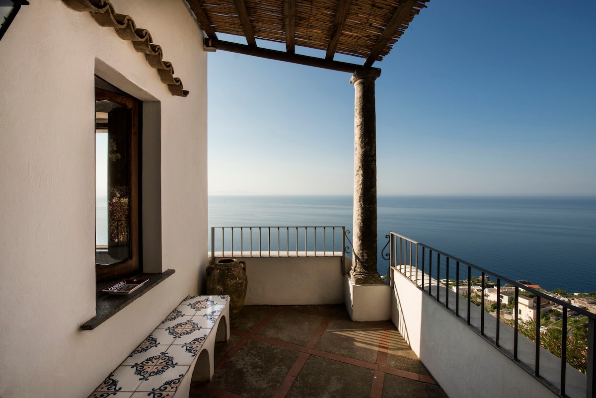 A shaded terrace offers a panoramic view of the calm sea, with a traditional tiled bench resting against the wall. A sturdy column supports the overhanging roof, creating a serene outdoor space perfect for relaxation.