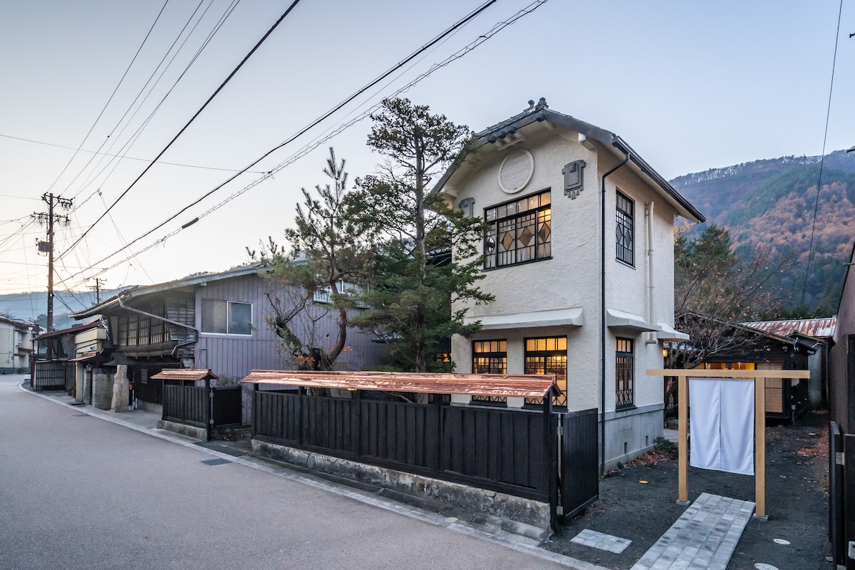The two-story building features a classic Japanese architectural style with white exterior walls and large windows framed in black. Surrounding trees and a small entrance gate enhance the serene atmosphere. The street is quiet, lined with traditional structures reflecting the town's historical charm.