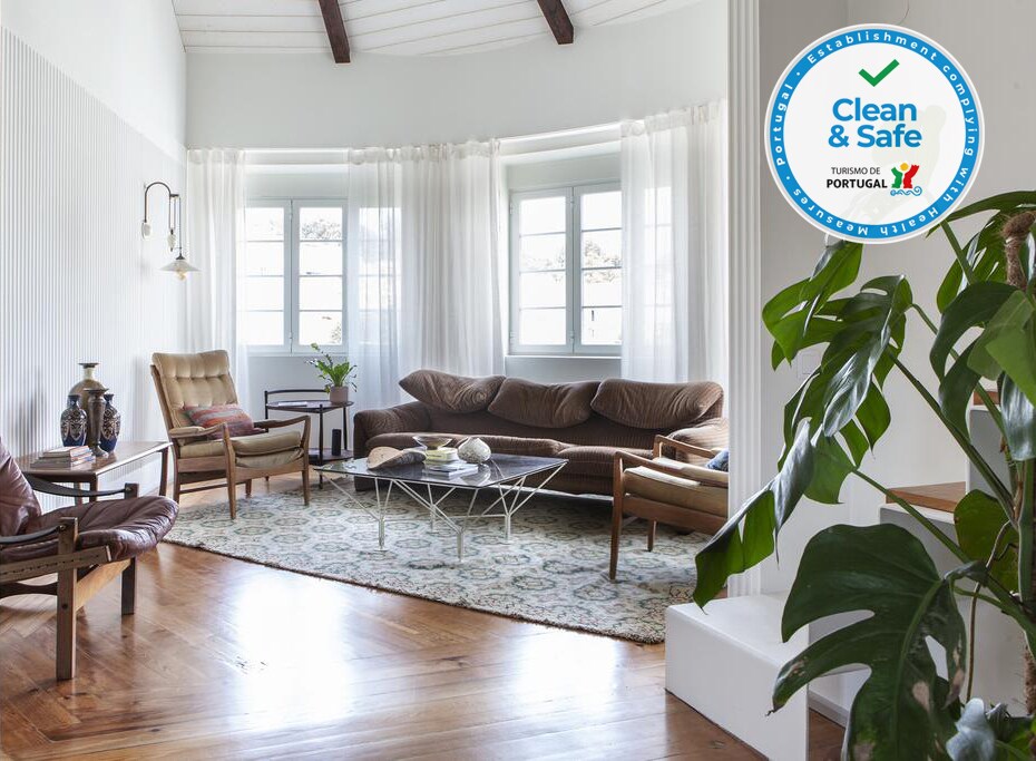 A multi-part living area features large windows allowing natural light to fill the space. A relaxed seating arrangement includes a leather sofa and wooden chairs, arranged around a coffee table on a textured rug. Light-colored curtains provide a soft ambiance.