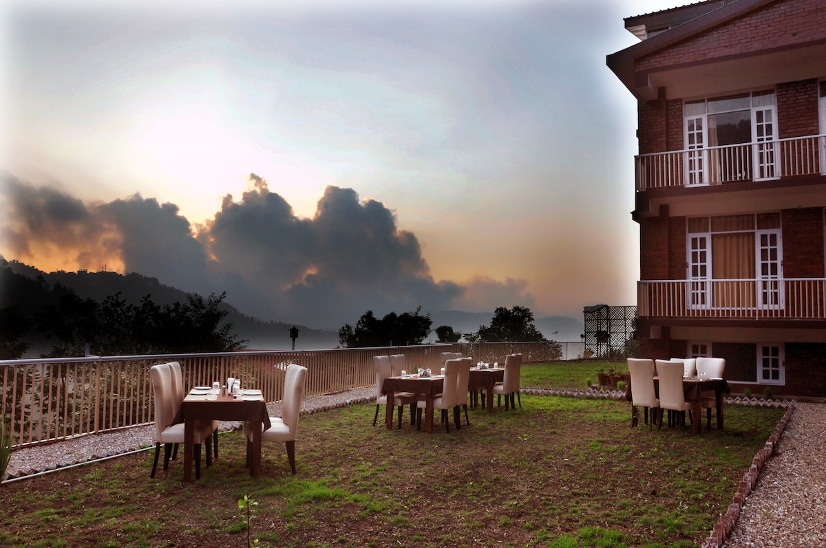 An outdoor dining area is set with wooden tables and cream-colored chairs, overlooking a hillside at sunset. The sky features soft clouds illuminated by warm hues, creating a serene backdrop for meals al fresco.