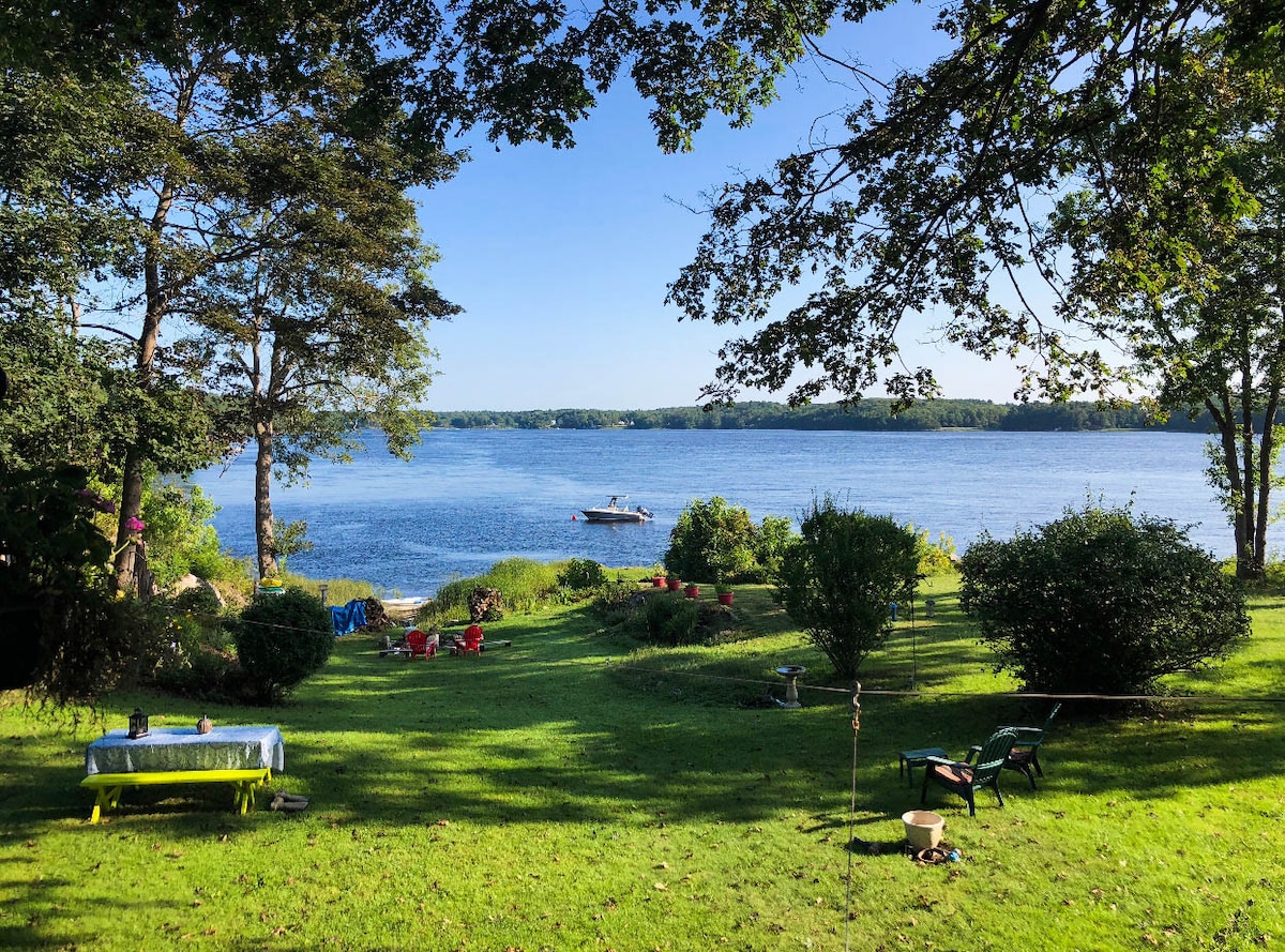 A serene view of the Kennebec River is framed by trees. The expansive lawn features scattered seating options, including colorful chairs and a dining table. A boat glides smoothly across the water, contributing to the peaceful ambiance of this natural setting.