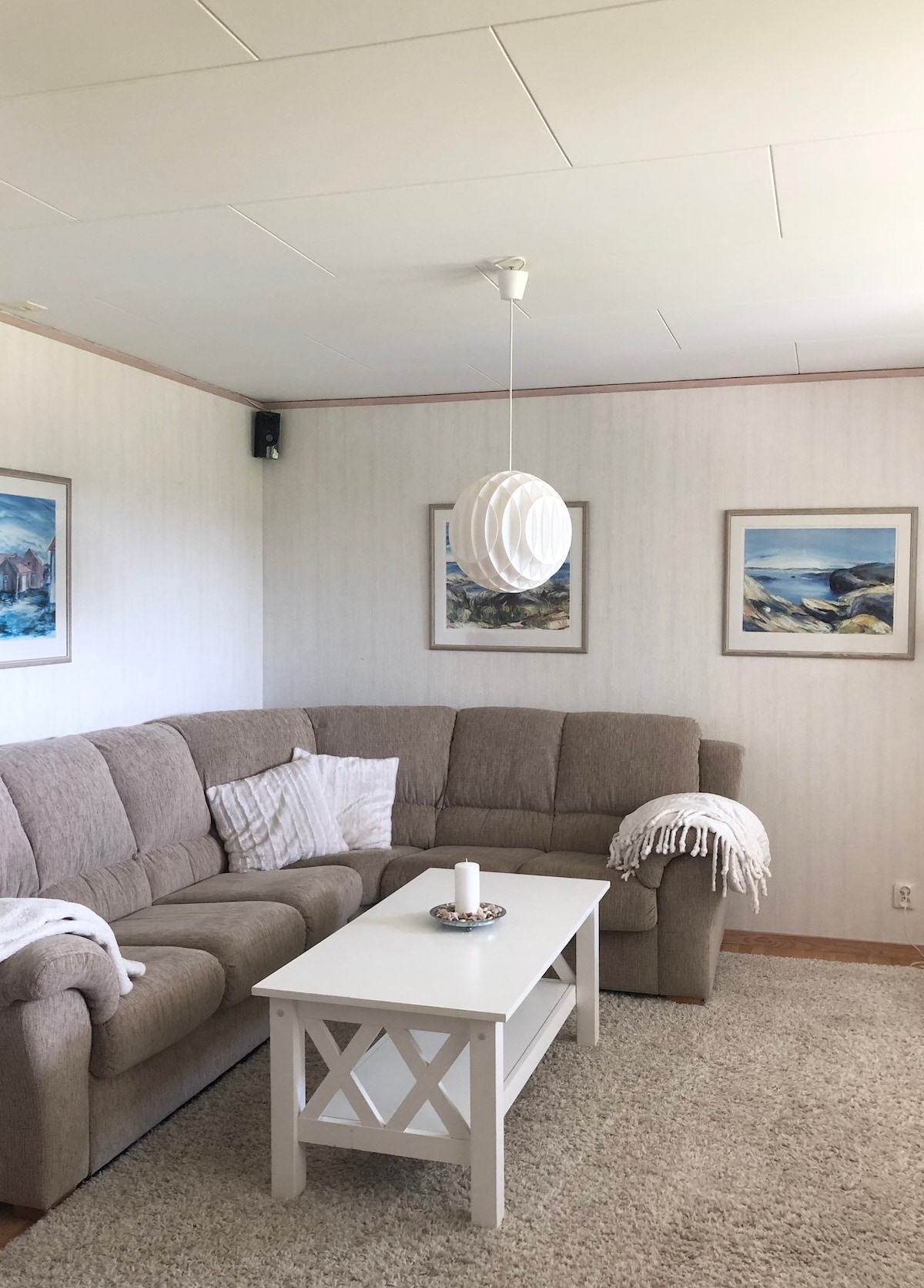 A cozy living area features a spacious sectional sofa in soft tones, complemented by two framed landscape artworks on the walls. A white coffee table sits at the center, adorned with a candle. A light fixture hangs from the ceiling above the seating.