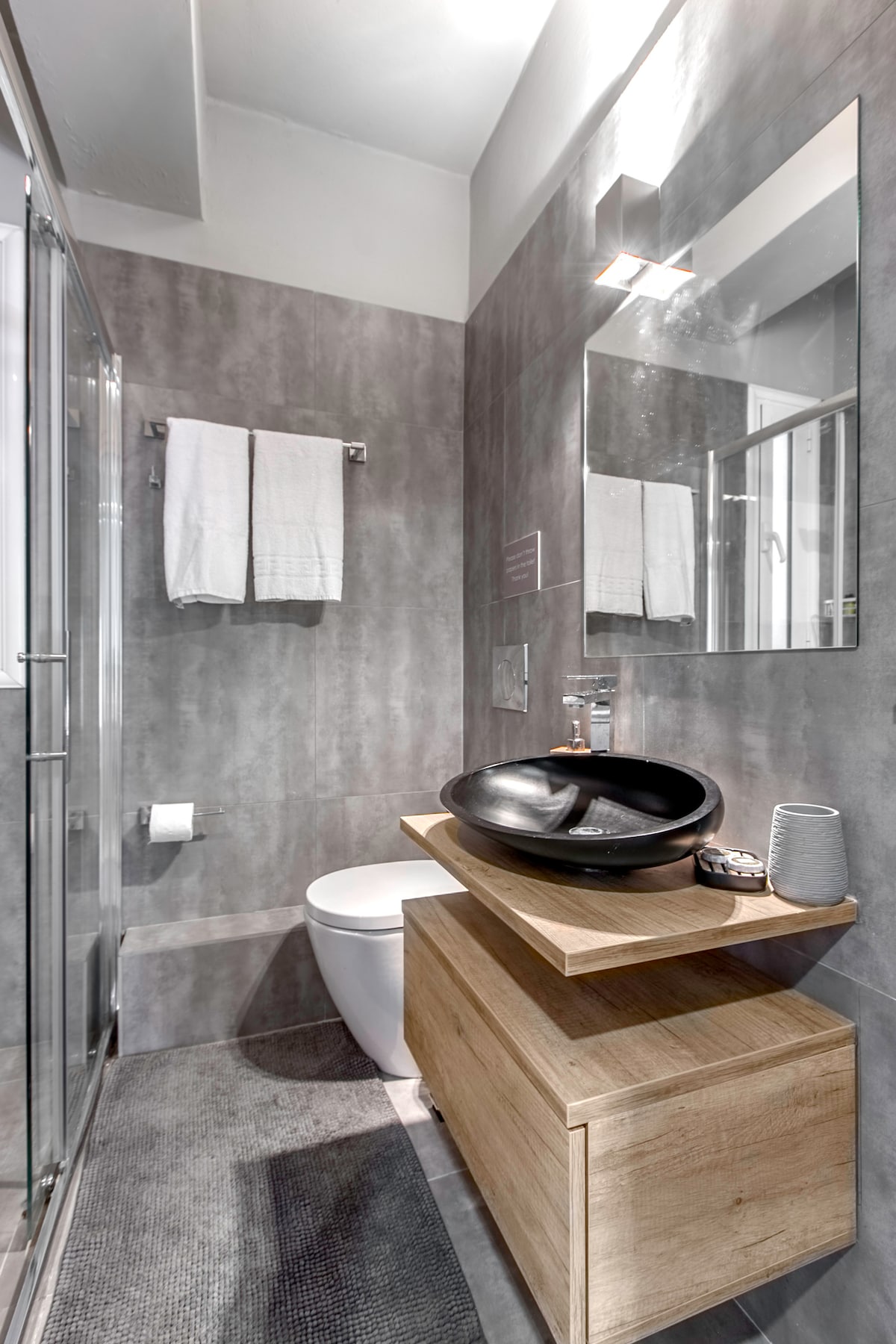 A modern bathroom is presented with sleek grey walls and lighting. A wooden vanity supports a round black sink and stylish accessories. Two white towels are neatly hung, and a large mirror reflects the tasteful design. A transparent shower enclosure is visible at the far end.