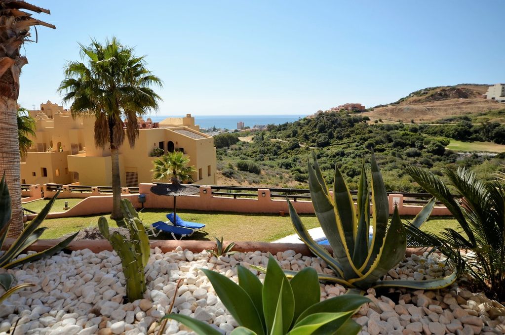 A vibrant garden area is presented, featuring a variety of tropical plants and neatly arranged white stones. In the background, a panoramic view reveals rolling hills and the sea in the distance under a clear blue sky.