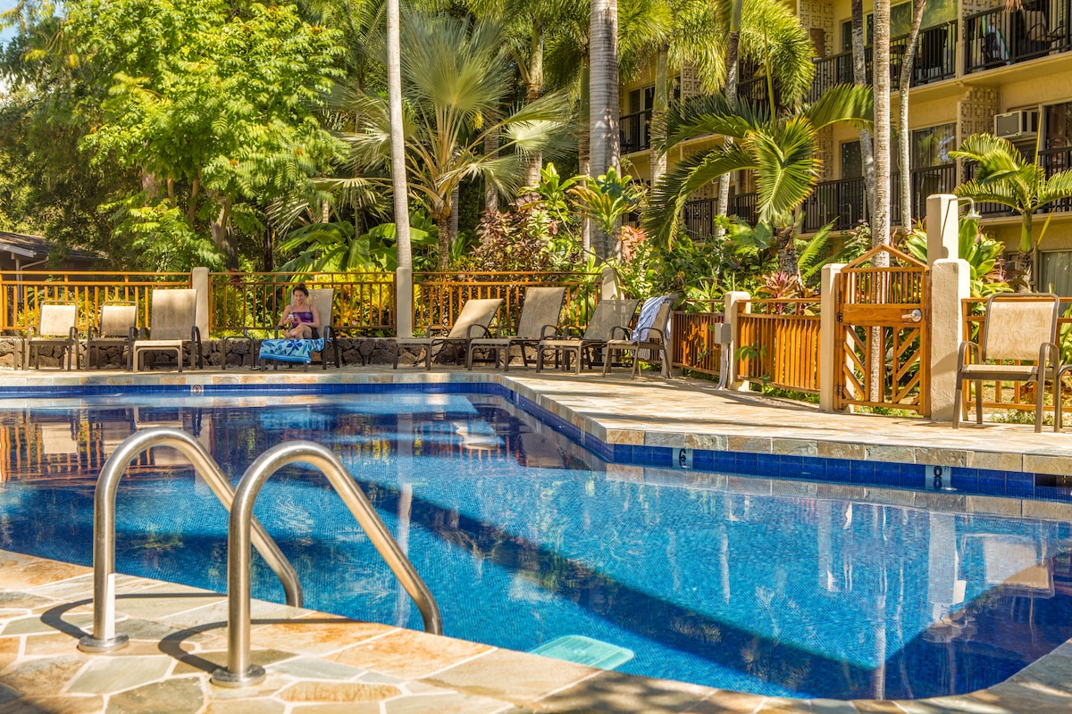 A serene pool area is surrounded by lush tropical landscaping and palm trees. Comfortable lounge chairs line the pool deck, inviting relaxation under the sun. The clear blue water of the pool reflects a bright sky, creating a tranquil atmosphere for guests.