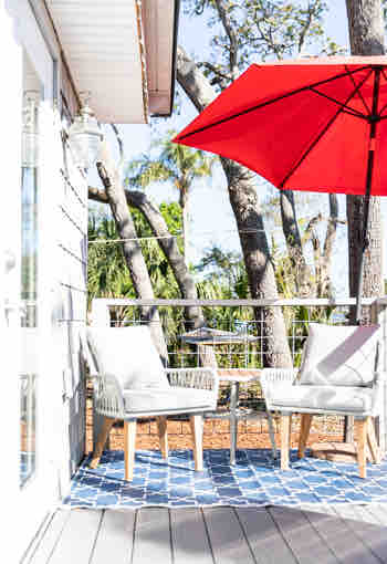 A small outdoor seating area is presented with two light-colored chairs on a patterned blue rug. A large red umbrella provides shade, while surrounding trees offer a natural backdrop, contributing to an inviting space for relaxation.