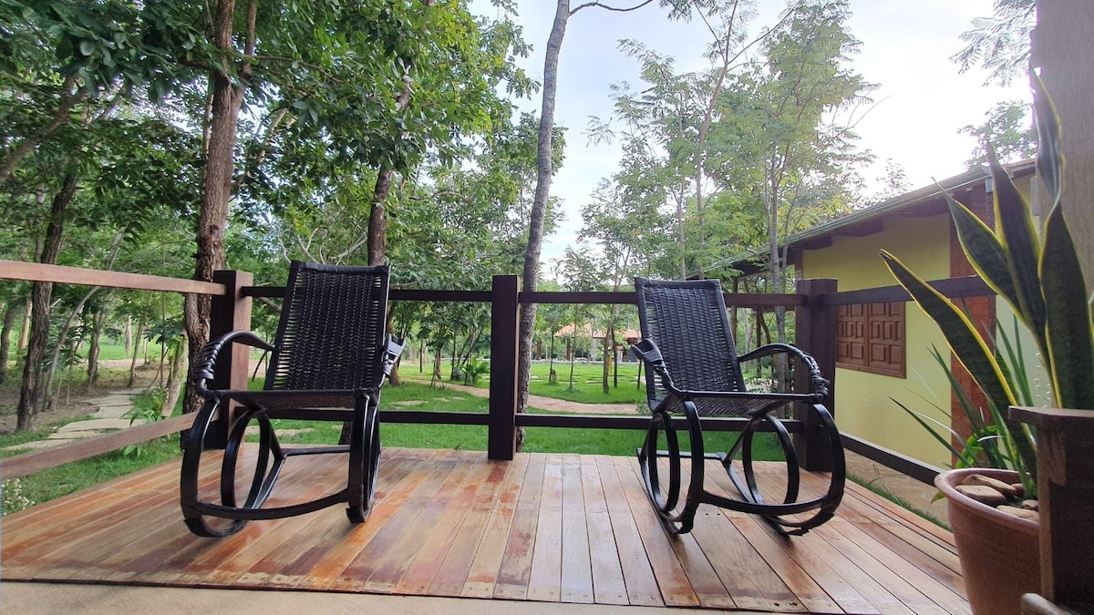 Two rocking chairs are positioned on a wooden deck, surrounded by lush greenery. Beyond the railing, a spacious lawn is visible, dotted with trees and well-maintained landscaping, providing a serene outdoor space for relaxation.