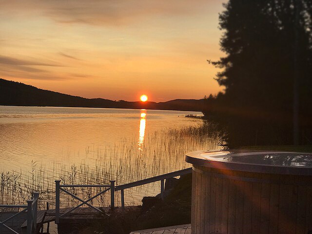 A serene sunset over Galasjön is visible, with the sun dipping toward the horizon. The reflection glimmers across the water, surrounded by tall grasses and distant silhouettes of trees. A wooden hot tub is positioned in the foreground, inviting relaxation with a scenic view.