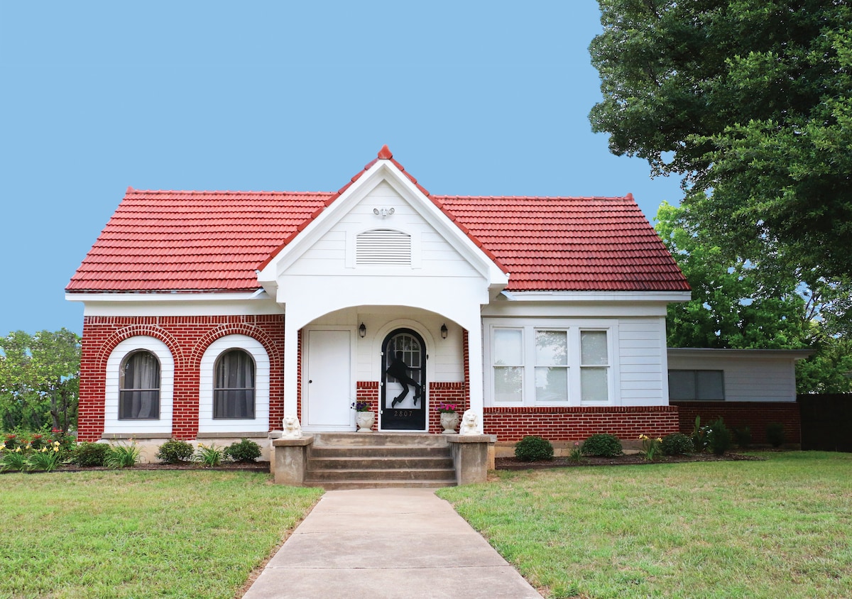 The historic home, accented by a bright red roof, features a blend of white siding and red brick. A set of steps leads to the entrance, framed by arched windows. Lush greenery surrounds the property, enhancing its charming curb appeal.