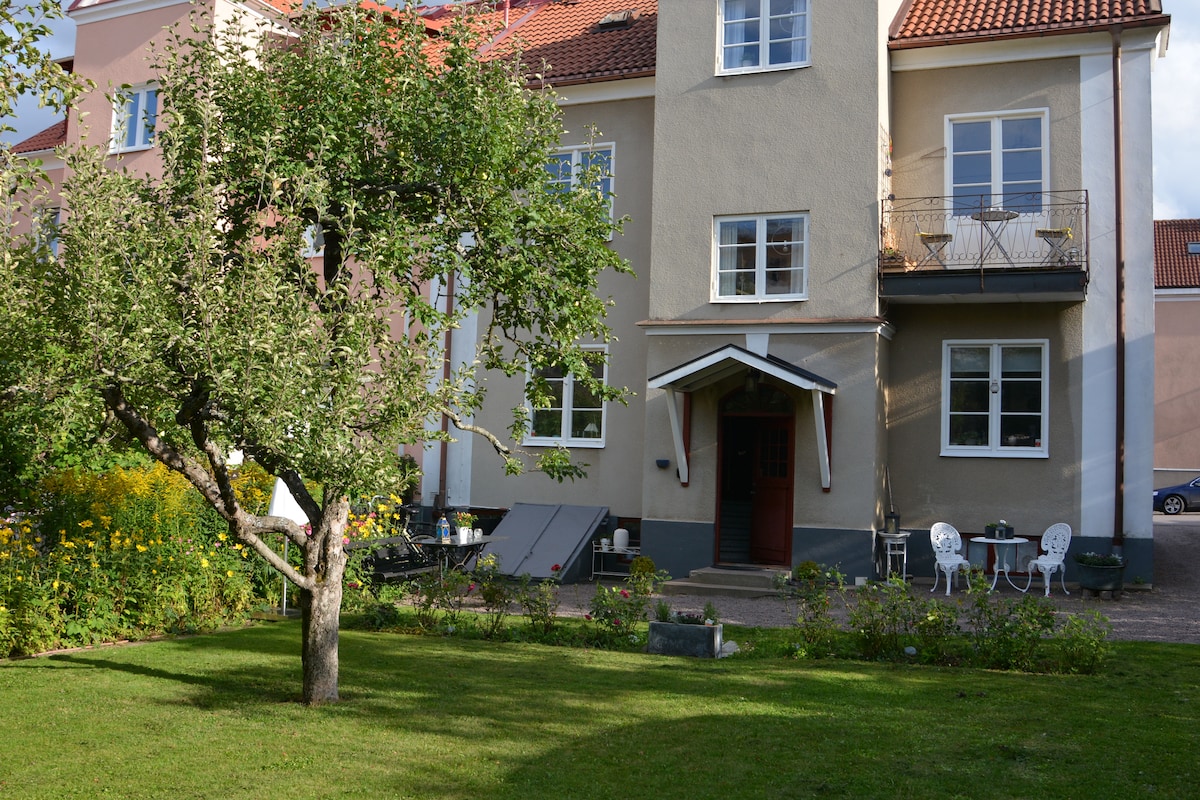 A charming building is surrounded by a peaceful garden featuring a lush lawn and an apple tree. Outdoor seating for two is positioned near the entrance, complemented by vibrant flower beds that enhance the tranquil atmosphere of the setting.