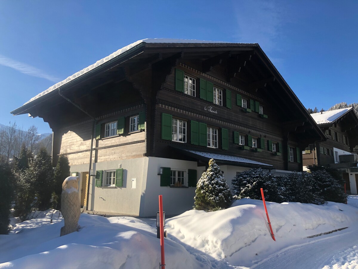 A charming chalet is depicted with traditional wooden architecture, accented by green shutters. Snow blankets the ground and the roof, creating a serene winter scene. Tall trees are visible nearby, enhancing the peaceful surroundings.