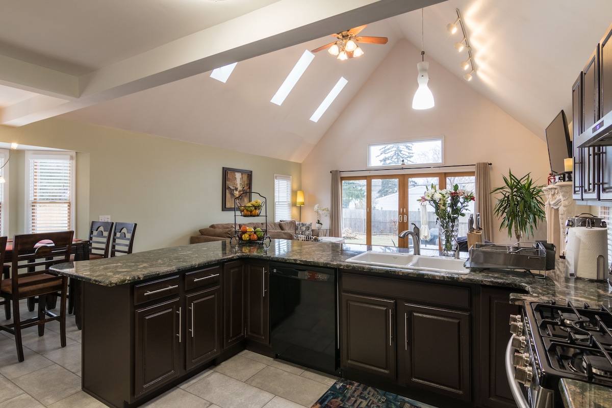 A spacious kitchen area is showcased with dark cabinetry and a large granite countertop. Natural light filters through skylights and large windows, illuminating the open layout. A dining set and relaxing seating area can be seen in the background, enhancing the inviting environment.