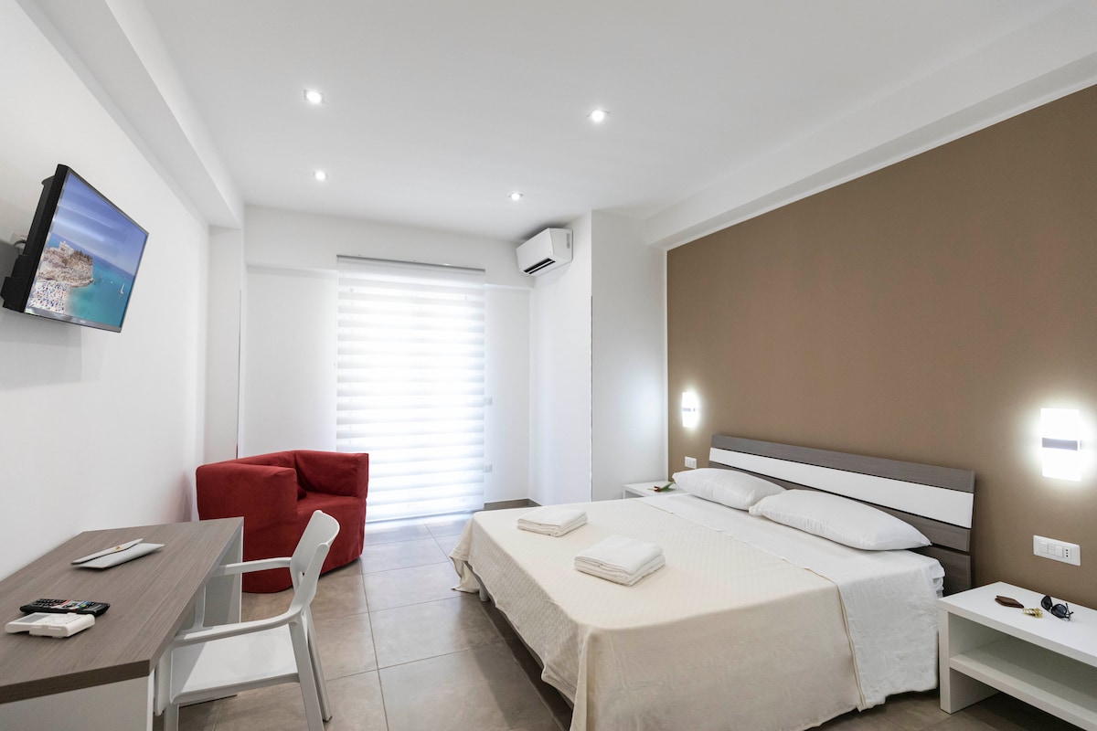 A modern bedroom features a spacious bed with neatly arranged linens. A cozy chair and desk are positioned against the wall, while large windows with soft blinds allow natural light to filter in. A wall-mounted television provides entertainment options.