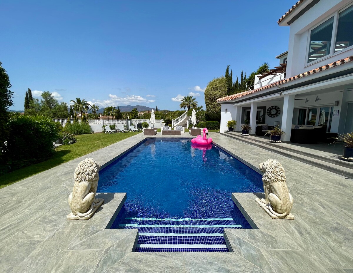 A private swimming pool is framed by elegant stone lions at each end. The water's deep blue hue reflects the clear sky above. Sun loungers rest nearby, while palm trees and lush greenery create a tranquil backdrop. A pink inflatable float adds a whimsical touch.