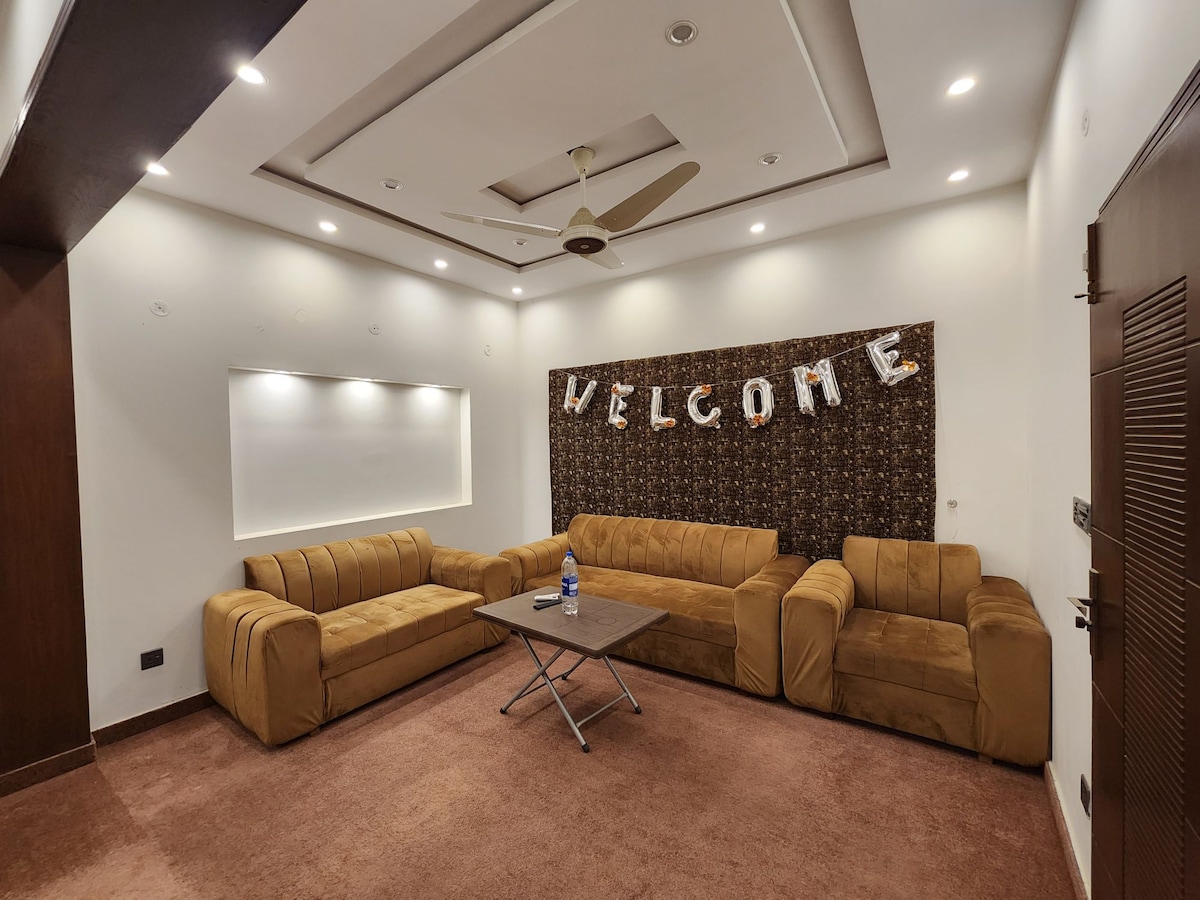 A spacious living area is furnished with plush, brown seating arranged in a conversational layout. A coffee table sits at the center, while a warm 'WELCOME' sign is displayed prominently on the wall, complemented by soft lighting from recessed fixtures.