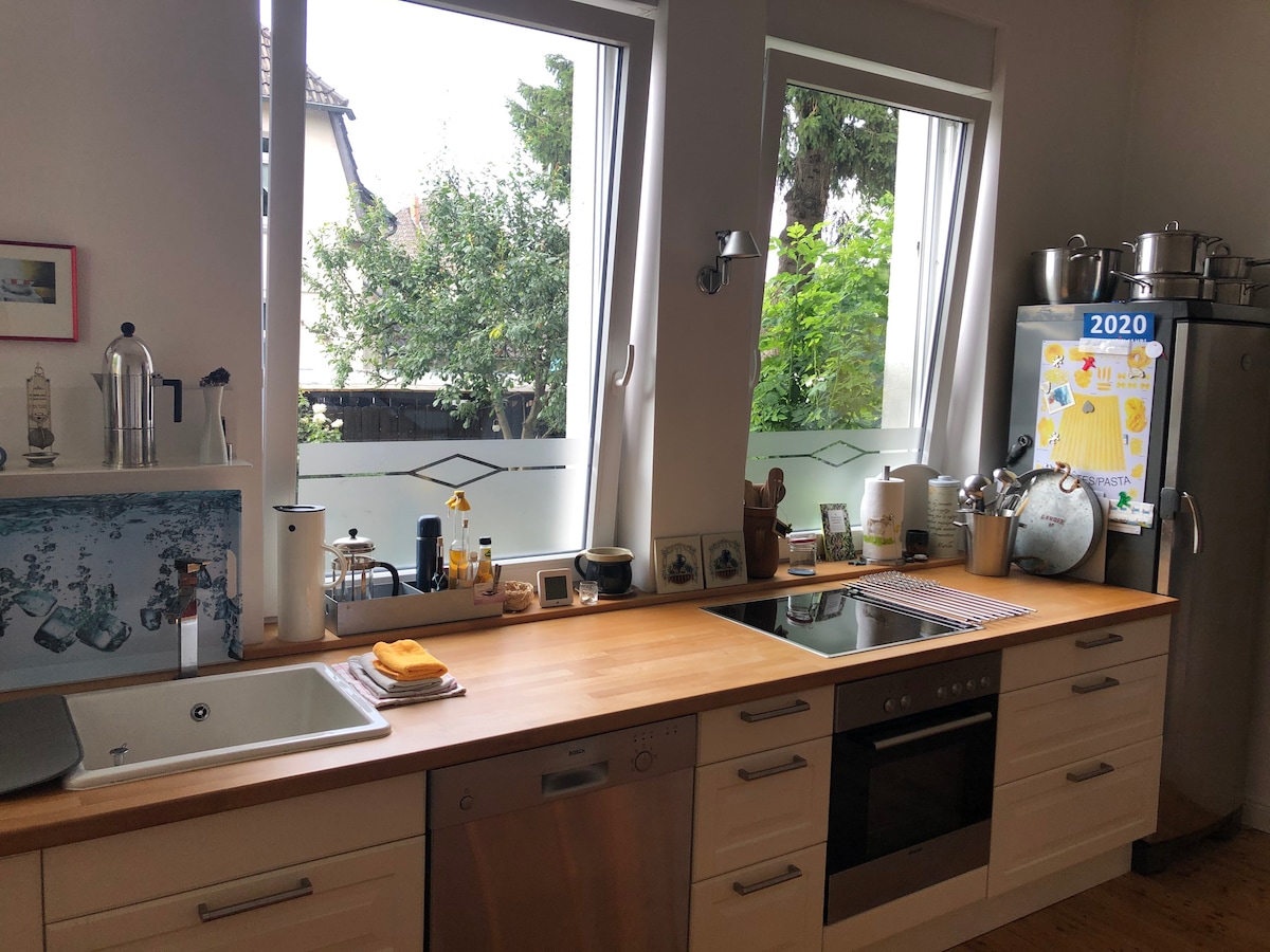 The kitchen features modern cabinetry and a wooden countertop. Cooking appliances, including an oven and stove, are visible alongside various kitchen tools. Natural light enters through large windows, providing views of greenery outside. Decorative elements, including a globe and artwork, are displayed on the counters.