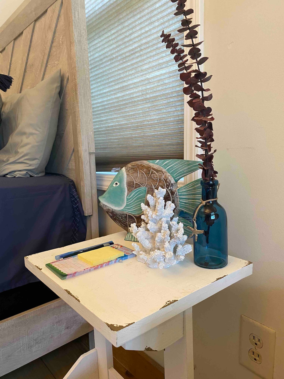 A bedside table is positioned next to a spacious bed, featuring a decorative turquoise fish and a textured coral sculpture. A blue glass bottle holds a slender arrangement of dried eucalyptus, while colorful notepads and pens are neatly placed on the table.