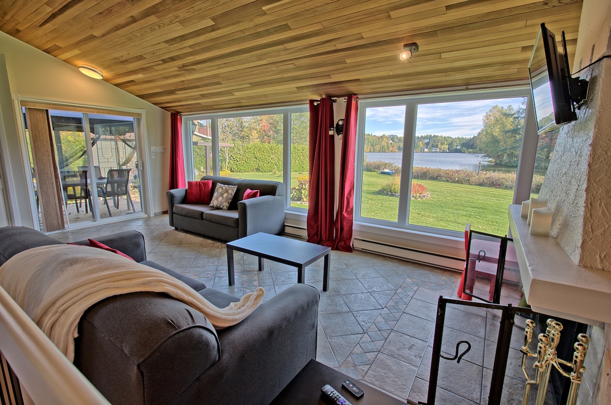 A spacious living area features comfortable gray sofas and a coffee table, with large windows offering a view of the lake and surrounding nature. Red curtains frame the windows, enhancing the serene ambiance. A fireplace is positioned to the side, adding to the cozy setting.