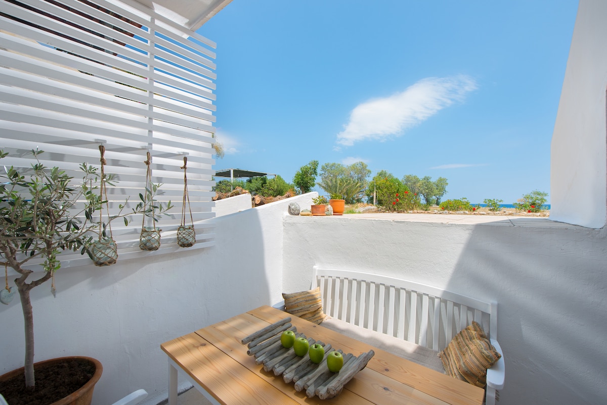 A bright outdoor terrace is shown, featuring a wooden table set with a decorative centerpiece of green apples. A white bench with soft cushions provides seating, while potted plants add greenery. The sky above is clear and blue, with distant trees visible in the background.