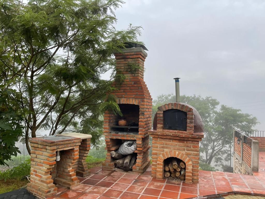 A rustic outdoor cooking area features a brick oven and barbecue grill, surrounded by greenery. The space includes a stone countertop and neatly stacked firewood, with a misty landscape in the background, creating a serene and inviting setting for outdoor dining.