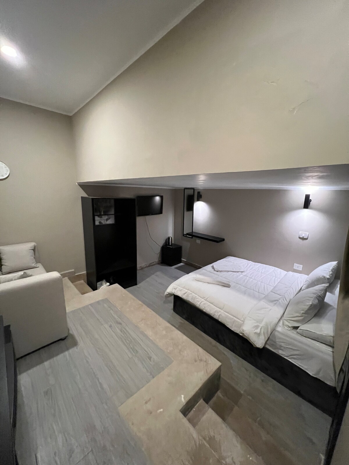 A compact studio features a king bed with crisp white bedding, adjacent to a modern seating area with a light-colored couch. A small entertainment unit and television are mounted on the wall, providing convenient access to media. Soft lighting creates a relaxed ambiance.