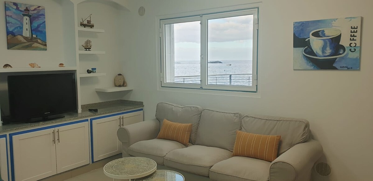 A comfortable seating area features a light-colored couch with striped cushions, positioned in front of a large window offering views of the sea. A flat-screen TV rests against the wall, while decorative elements include shelf displays and artwork that enhance the coastal ambiance.