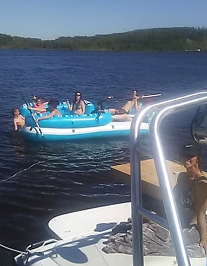 A group of individuals relaxes on an inflatable raft floating on calm water, surrounded by greenery and distant hills. The scene is sunny, with clear skies above, creating a tranquil atmosphere. A boating platform is visible in the background, adding to the recreational setting.