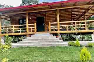 A wooden cabin with a red roof is surrounded by greenery. The entrance is accessed by a set of stone steps, leading to a spacious porch with wooden railings. Lush grass and small shrubs are present in the foreground, creating a serene natural setting.