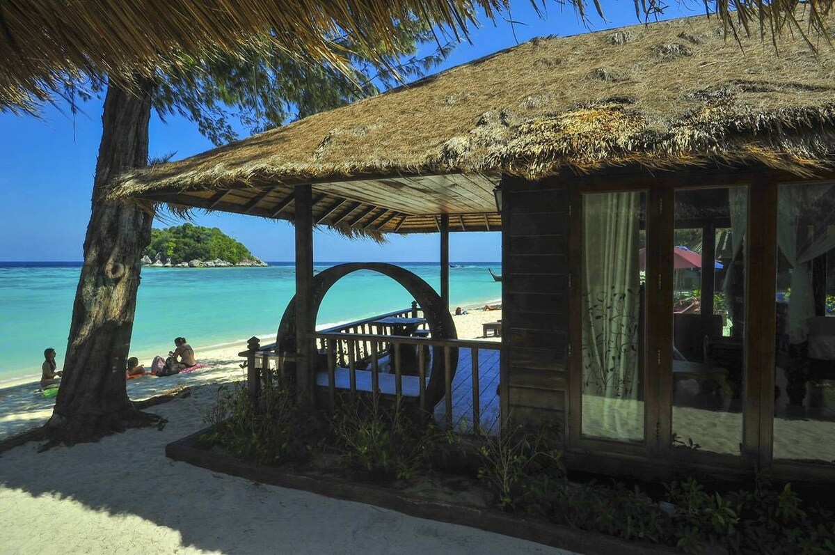 A beachfront bungalow featuring a thatched roof is positioned near soft white sand. Palm trees provide shade, with clear turquoise waters visible in the background. Guests are seen enjoying the beach, highlighting the tranquil environment.