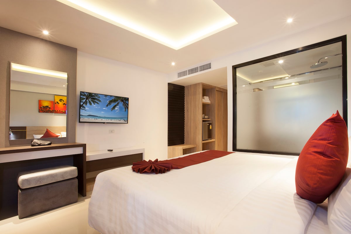 A modern bedroom features a spacious bed with crisp white linens and a decorative red pillow. A large wall-mounted television is positioned across from the bed. A sleek vanity area with a mirror is seen, along with a wooden wardrobe and frosted glass accents enhancing the room's contemporary design.