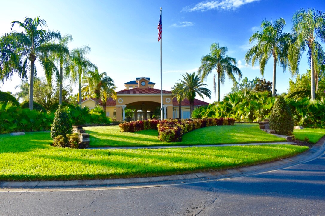 The entrance of the resort is framed by lush palm trees and well-maintained landscaping. A manicured lawn surrounds the circular driveway, leading to a welcoming building featuring a flagpole with an American flag. Bright blue skies provide a vibrant backdrop.