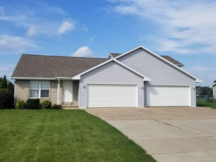 A single-story duplex is shown with a light gray exterior and a two-stall garage. The neatly paved driveway leads to the garage doors, while a well-maintained lawn with green grass and slight landscaping is visible in the foreground under a partly cloudy sky.