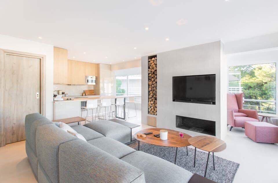 A contemporary open living area features a spacious gray sofa with a textured rug beneath. Light wooden cabinetry complements the modern kitchen, while a television is mounted on the wall. A cozy seating area is created by a pink armchair near the window, inviting natural light.